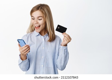 Excited Blond Teen Girl, Child Showing Credit Card Unior, Looking Happy At Smartphone App Screen, Standing Over White Background, Bank Account For Children Concept