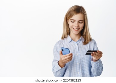 Excited Blond Teen Girl, Child Showing Credit Card Unior, Looking Happy At Smartphone App Screen, Standing Over White Background, Bank Account For Children Concept.