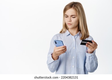 Excited Blond Teen Girl, Child Showing Credit Card Unior, Looking Happy At Smartphone App Screen, Standing Over White Background, Bank Account For Children Concept.