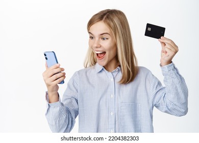 Excited Blond Teen Girl, Child Showing Credit Card Unior, Looking Happy At Smartphone App Screen, Standing Over White Background, Bank Account For Children Concept.