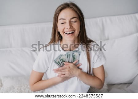 Excited blonde caucasian girl broad smiling eyes closed presses banknotes to chest, got salary, sits on bed. Happy Swedish young woman received money, celebrates. Prize, lottery, winner. Wage. Cash