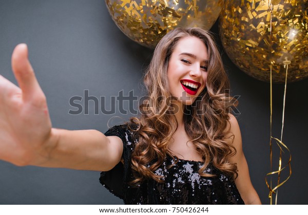 お祭りの間 暗い背景に自撮りをする赤い唇をした興奮した金髪の女の子 パーティー風船の前にポーズをとる黒いドレスを着た ヨーロッパ人の喜ばしい女性モデルの室内ポートレート の写真素材 今すぐ編集