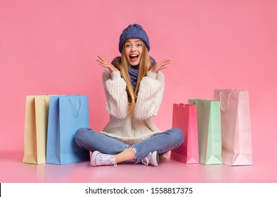 Excited blonde girl in winter blue set sitting on floor among colorful shopping bags over pink background, raising hands in emotional gesture, copy space