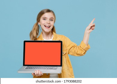 Excited blonde little kid girl 12-13 years old in yellow jacket isolated on blue background. Childhood lifestyle concept. Hold laptop pc computer with blank empty screen point index finger aside up