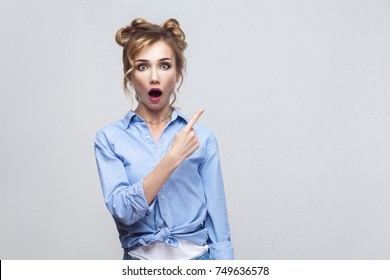 Excited blonde woman pointing finger sideways, raising eyebrows and keeping mouth wide opened, showing something surprising on grey copy space wall. Indoor, studio shot