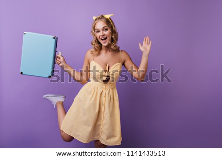Excited blonde woman in yellow dress waiting for vacation. Studio portrait of emotional curly girl funny dancing on purple background.