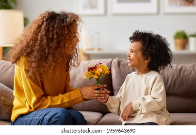 Excited Boy Little Son Congratulating His Mom Happy Mixed Race Woman With Mothers Day And Giving Her Flower Bouquet, They Smiling While Sitting On Sofa At Home. Family Holidays Concept