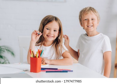 Excited Boy Standing Near Sister Sitting At Table Near Colorful Felt Pens