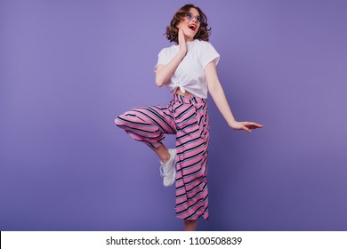 Excited brown-haired girl in stylish attire fooling around during indoor photoshoot on purple background. Debonair european woman in white t-shirt jumping in studio.