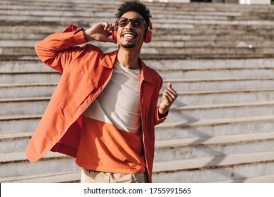 Excited brunet charming man in orange jacket, colorful t-shirt and sunglasses sings, sincerely smiles and listens to music in headphones outside.