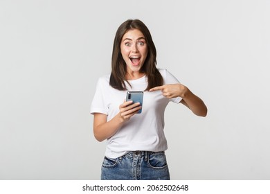 Excited brunette girl smiling amazed and pointing at smartphone screen