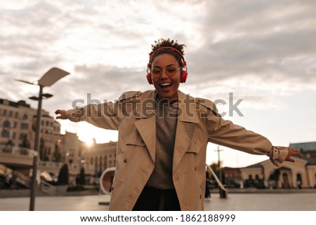 Excited brunette woman in beige trench coat listens to music in red headphones and dances in great mood outdoors during sunset.