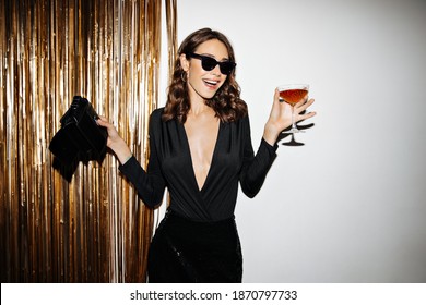 Excited Brunette Woman In Sunglasses Has Fun And Holds Cocktail Glass. Joyful Lady In Festive Dress Dances Near Wall With Gold Confetti.