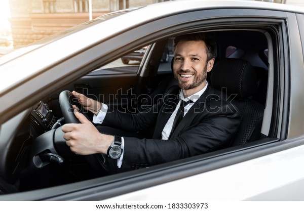 Excited businessman driving car to airport, going\
on business trip, smiling at camera through luxury car window, side\
view, copy space. Happy man in suit going to business meeting in\
the morning