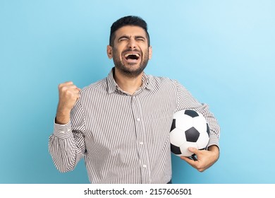 Excited businessman screaming widely opening mouth, celebrating victory of favourite football team, holding soccer ball in hands, wearing striped shirt. Indoor studio shot isolated on blue background.