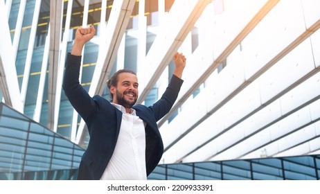 Excited Businessman In Suit Celebrating Victory Arms Raised Up. Hispanic Male Business Person Winner Pose. Best Deal Ever. Business Man Expressing Positivity Standing Outdoors Office Building