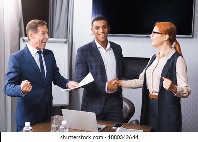Excited businessman with yes gesture standing near multicultural colleagues shaking hands with each other in meeting room - Shutterstock ID 1880342644