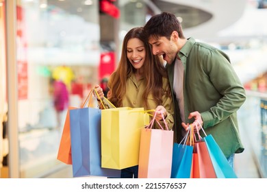 Excited Buyers Couple Looking Inside Shopping Bags Buying New Clothes Together During Black Friday Sales Season Standing In Modern Hypermarket Indoor. Discounts Offer Concept