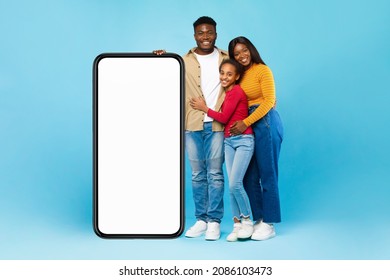 Excited Casual Black Family Leaning On Big Smartphone With White Empty Cell Screen, Cheerful Mom, Dad And Daughter Hugging, Recommending New App Or Website, Standing At Blue Wall, Mock Up, Full Body