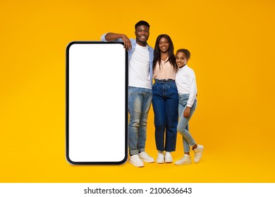 Excited Casual Black Family Leaning On Big Smartphone With White Empty Cell Screen, Cheerful Mom, Dad And Daughter Recommend New Website App, Standing At Yellow Orange Wall, Mock Up, Full Body Length