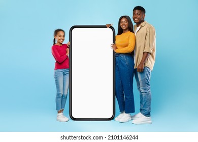 Excited Casual Black Family Leaning On Big Smartphone With White Empty Cell Screen, Cheerful Mom, Dad And Daughter Recommend New Website App, Standing At Blue Studio Wall, Mock Up, Full Body Length