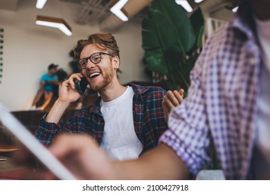 Excited Caucasian Businessman Talking On Smartphone While His Cropped Male Colleague Using Mobile Phone. Business Cooperation And Teamwork. Modern Successful Men. Young Guys Working In Office Space