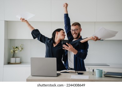 Excited caucasian couple after calculate their year earnings, celebrating successful business at new home using calculator, laptop. Woman raises hands in winner gesture holds papers. Financial freedom