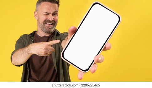 Excited Caucasian Mature Man Holding Big Smartphone With White Blank Screen In Hand, Showing Close To Camera And Pointing At Device. Big Gadget With Empty Free Space For Mock Up, Banner Yellow Wall.