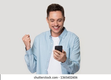 Excited Caucasian Millennial Man Isolated On Grey Studio Background Look At Smartphone Happy Get Good Text Message, Smiling Male Client Use Cellular Read Great News, Win Lottery On Mobile App Online