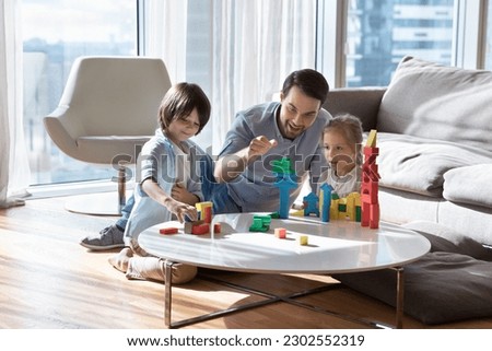 Excited cheerful dad enjoying leisure playtime with two little children, stacking toy tower with kids, teaching son and daughter to construct castle, enjoying parenthood, weekend