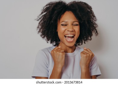 Excited cheerful dark skinned girl clenches fists while closed her eyes exclaims with victory or willing to win, excited female dressed in casual wear shouts yeah, isolated on white