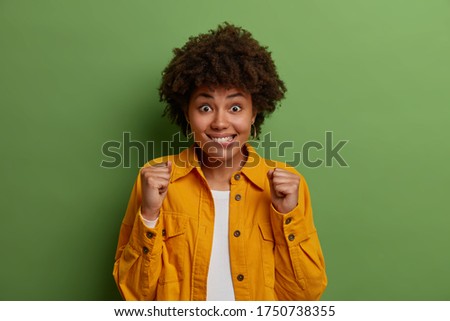 Excited cheerful dark skinned woman roots for favorite team, clenches fists and bites lips with great anticipation of win, cheers and hopes for good deal, rejoices winning in lottery, poses indoor