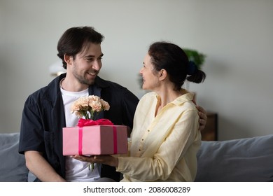 Excited cheerful middle aged mom receiving surprise gift and flower bunch from happy grateful adult son, celebrating birthday, mothers day, 8 march at home, holding present box, bouquet