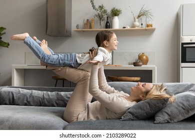 Excited Cheerful Mom Lifting Daughter Kid, Holding Little Girl In Air With Open Flying Hands, Playing Airplane, Resting On Soft Couch, Laughing, Practicing Acroyoga With Child. Family Active Games