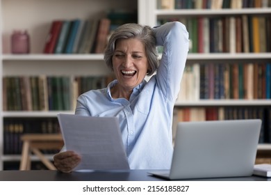 Excited Cheerful Senior Business Lady Reading Document At Workplace With Laptop, Reviewing Paper, Laughing. Happy Older Retired Woman Satisfied With Insurance Agreement Terms