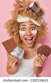 Excited Cheerful Woman Has Comb Stuck In Curly Hair Exclaims Loudly Wears Blindfold And T Shirt Stares Impressed Applies Beauty Patches Under Eyes To Remove Puffiness Holds Bar Of Chocolate And Cookie