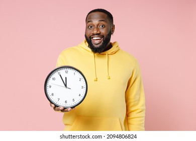 Excited Cheerful Young African American Man 20s Wearing Casual Yellow Streetwear Hoodie Standing Holding In Hand Clock Looking Camera Isolated On Pastel Pink Color Wall Background Studio Portrait