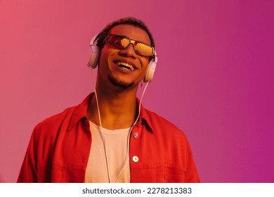 Excited cheerful young african man in sunglasses listening music with headphones isolated over multicolored neon studio background - Powered by Shutterstock