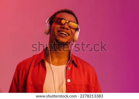 Excited cheerful young african man in sunglasses listening music with headphones isolated over multicolored neon studio background