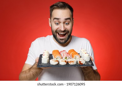 Excited cheerful young bearded man 20s wearing casual white t-shirt looking on makizushi sushi roll served on black plate traditional japanese food isolated on red color background studio portrait
