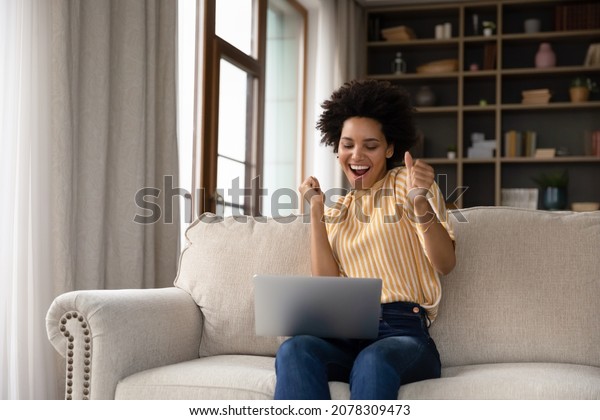 Excited cheerful young Black woman using laptop\
computer on sofa at home, getting good news, feeling joy, dancing\
with hands, singing, laughing, making winner gesture, happy to win\
prize