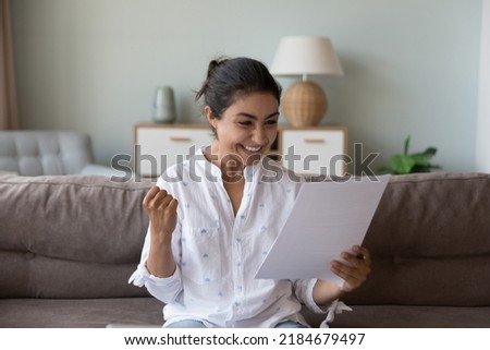 Excited cheerful young Indian woman getting good news letter, reading paper document at home, making winner yes hand, feeling happy, smiling, laughing, celebrating, success, achieve