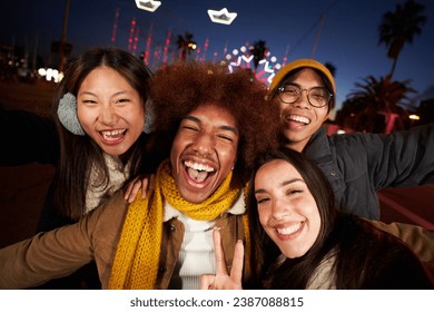 Excited Christmas selfie of multicultural young group friends. Four happy colleagues taking mobile photo looking smiling at camera standing together at winter night. Generation z people posing outdoor - Powered by Shutterstock