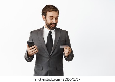 Excited Corporate Man Looking At His Credit Card While Using Mobile Phone App, Paying Online, Transfer Money Via Smartphone Application, White Background