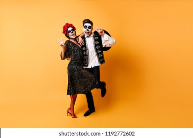 Excited Couple In Halloween Costumes Expressing Positive Emotions. Studio Full-length Portrait Of Funny Zombies Dancing On Bright Background.