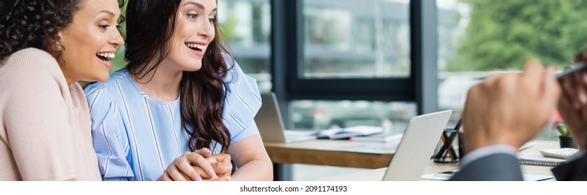 Excited Couple Of Interracial Lesbian Women Holding Hands While Looking At Laptop Near Blurred Realtor, Banner