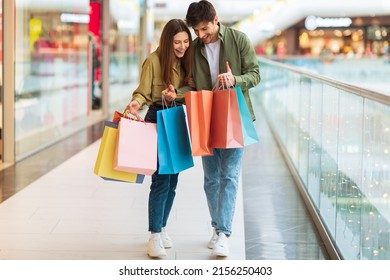 Excited Couple Looking Into Shopping Bags Enjoying Sales Weekend In Modern Mall, Buying New Clothes. Full Length Of Happy Buyers Standing In Hypermarket. Commerce Concept
