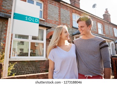 Excited Couple Standing Outside New Home With Sold Sign