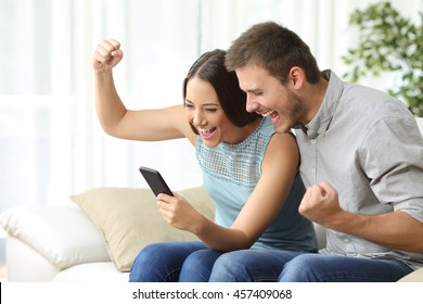 Excited Couple Watching Media Content Together Using A Mobile Phone Sitting On A Couch In The Living Room Of A House