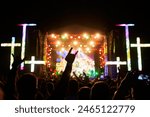 Excited crowd at night, illuminated by colorful stage lights at outdoor Christian rock fest. Fans raise hands, enjoy lively music, united in faith and celebration. Band performs live, energy high.